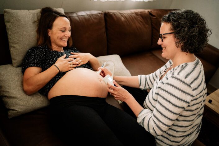 Schwangere freut sich bei Hausbesuch von Hebamme, die mit Bauch abhört