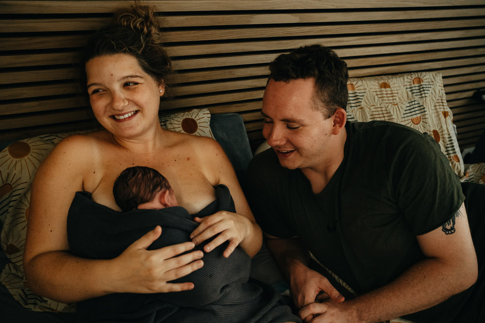 Vater blickt auf frisch Geborenes Baby im Arm der Mutter direkt bei Hausgeburt Reportage