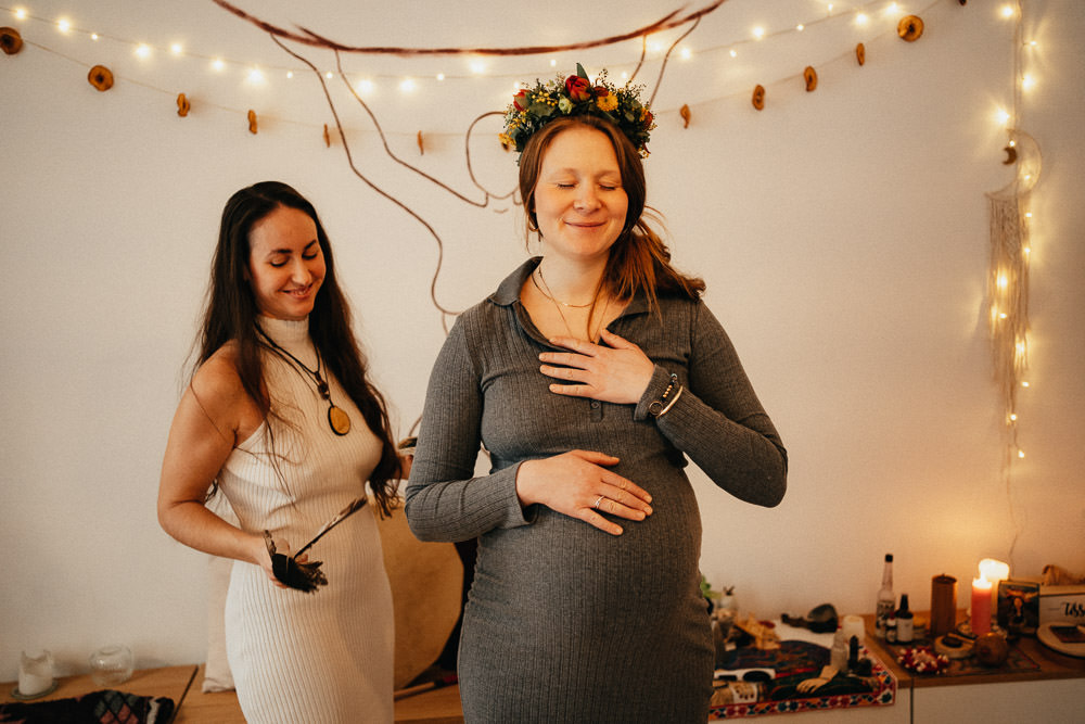 Frau räuchert schwangere Mutter beim Mothers Blessing und diese trägt eine Blumenkrone und hat die Hand auf dem Babybauch
