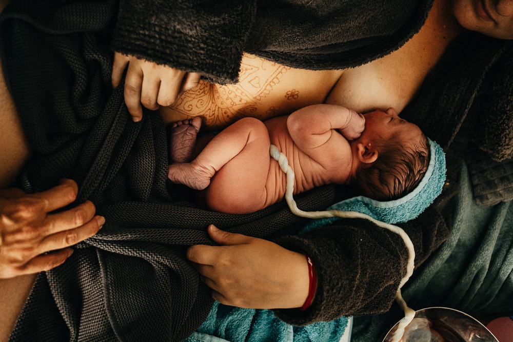 Stillfoto von frisch geborenem Baby bei einer Hausgeburt mit viel Haut und Falten auf dem Bauch der Mutter mit der Nabelschnur die noch zur Plazenta führt